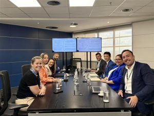Meryem Duygun and and Linh Nguyen sitting down in a boardroom at a large black wooden table with people from The Caixa Econômica Federal and 2 screens in the background