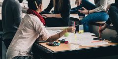 A group of people sit around a table which is covered in paper and pens. They appear to be in conversation, but we can't see the faces of any of the people.