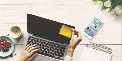 Two hands are place on a laptop. The right hand is putting a sticky note on the laptop screen. A phone next to the laptop has a number of social media apps visible.