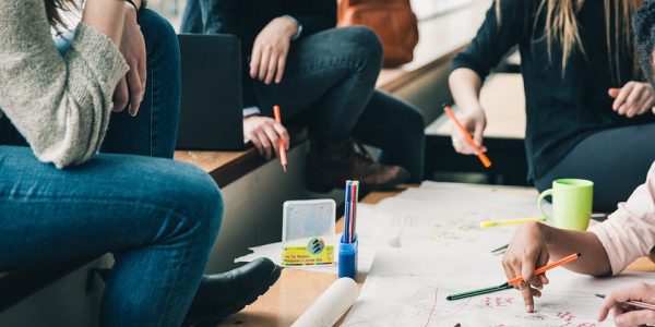 A group of people (faces not shown) are sat around a table with paper and pens, brainstorming.