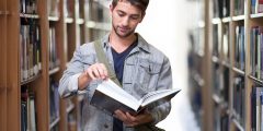 Male student in library surrounded by books is facing towards the camera whilst looking at an open book
