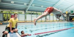 A male in red swimming trunks diving into a swimming pool with two other males watching