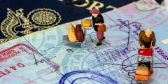 A passport, underneath an open passport showing stamps, with little figurines on top that are wheeling luggage trolleys.