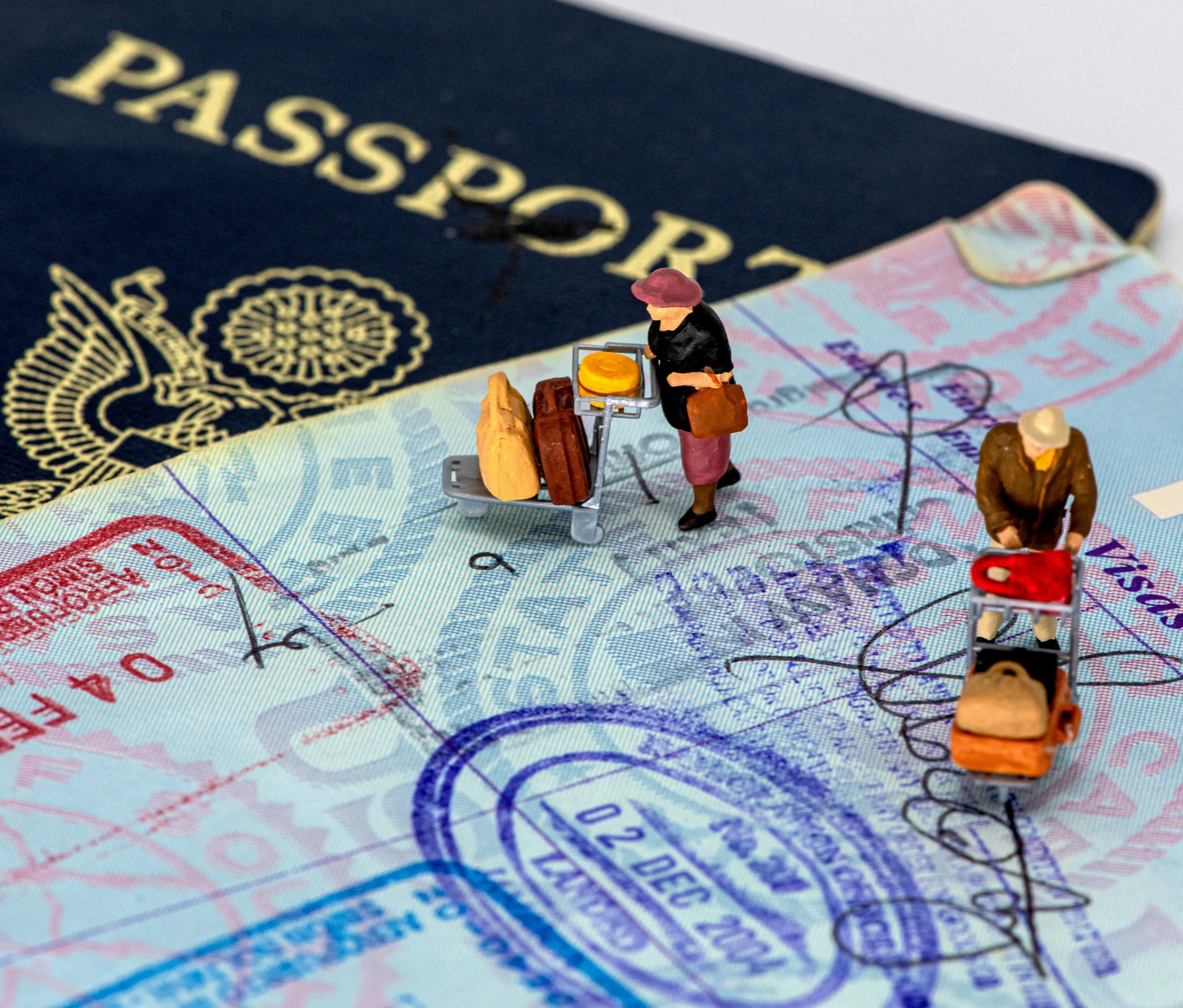 A passport, underneath an open passport showing stamps, with little figurines on top that are wheeling luggage trolleys.