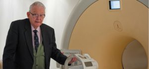 An elderly Mansfield, dressed in a suit, standing next to a modern MRI