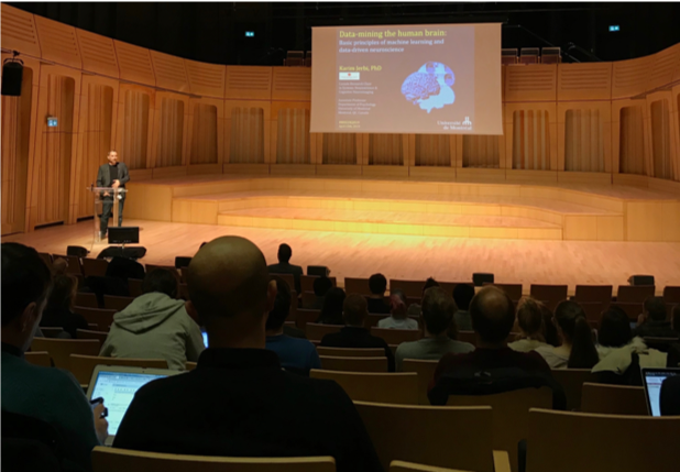view from lecture theatre audience of man at podium with projection behind of slide with image of a human brain