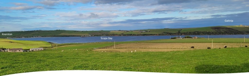 Colour photo of view over Scapa Bay. The foreground is green pasture land, the middle ground is the bay and beyond re sea cliffs topped with farm and heath land. The sky is blue with white and grey clouds stretching across it. Scapa bay is in the centre.
